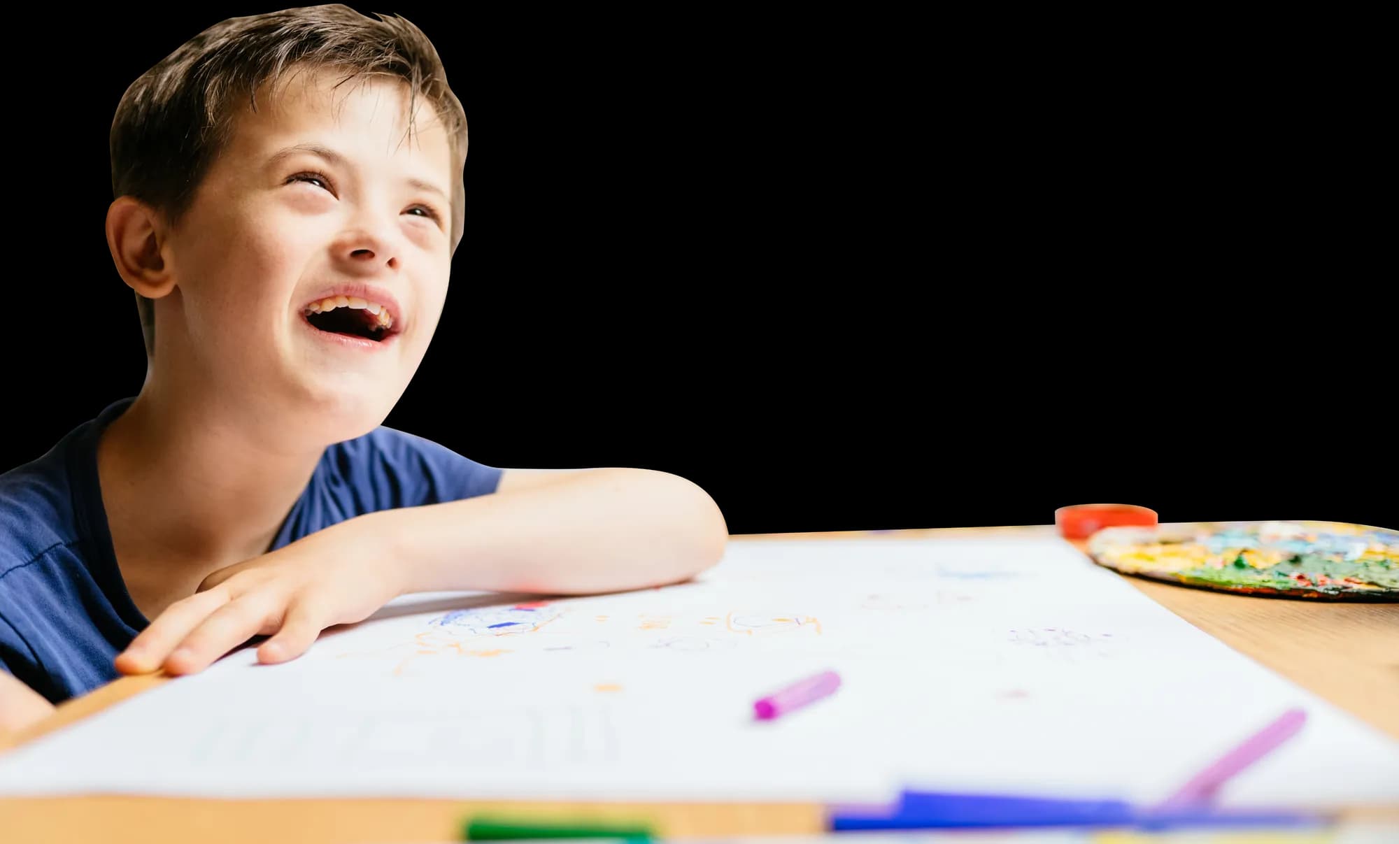 Smiling boy with Down syndrome leaning on an art table, drawing with crayons